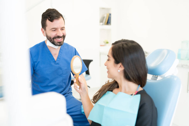 Usmívající se muž ortodontista při pohledu na ženu držící zrcadlo po ortodontické léčbě - Fotografie, Obrázek