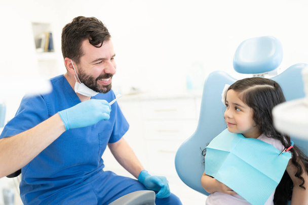 Маленькая латиноамериканка смотрит на стоматолога-мужчину с помощью зубной щетки в клинике - Фото, изображение
