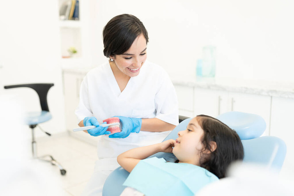 Magabiztos kaukázusi női fogorvos megmutatja egy kislánynak, hogyan kell megfelelően fogat mosni egy fogászati klinikán. - Fotó, kép