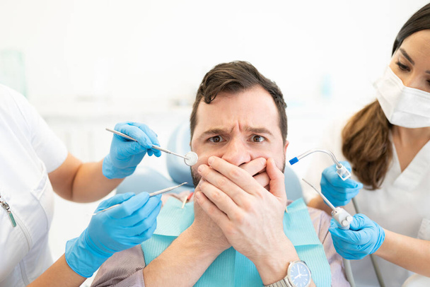 Spaventato uomo medio adulto che copre la bocca mentre il dentista tiene gli strumenti durante il trattamento - Foto, immagini
