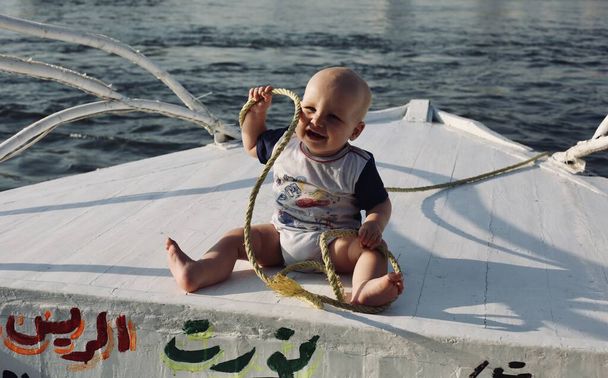 Bambino di 7 mesi seduto a poppa della vecchia barca bianca, nave piacere, sorriso e giocare con la corda durante il viaggio turistico al fiume Nilo al Cairo, Egitto  - Foto, immagini