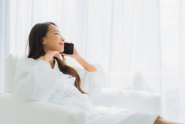 ポートレート美しい若いアジアの女性は、リビングルームのエリアでソファにスマートフォンを使用します - 写真・画像