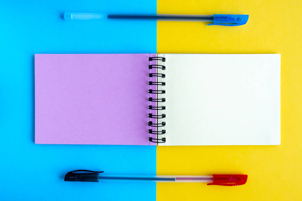 Auf gelb-blauem Hintergrund liegt ein Notizbuch mit mehrfarbigen Seiten und zwei Kugelschreibern. Draufsicht, flache Lage, Kopierraum. - Foto, Bild