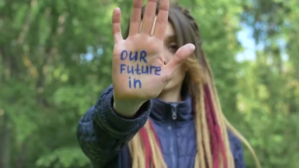 長いドレッドロックを持つ現代の深刻な女の子は書かれたスローガンで手を示しています緑の木の背景にあなたの手に私たちの未来。責任、気候変動の概念 - 映像、動画