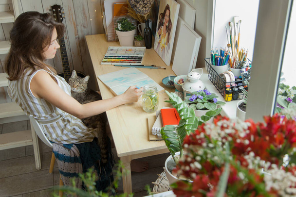 The girl artist sits at her workplace and paints portraits in watercolors. Finished works are hanging on the wall. She has a gray cat in her arms. On the table are brushes, paints, paper, tea and flowers. - Foto, Imagem