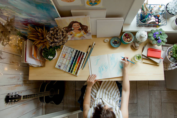 The girl artist sits at her workplace and paints portraits in watercolors. Finished works are hanging on the wall. She has a gray cat in her arms. On the table are brushes, paints, paper, tea and flowers. - Fotó, kép