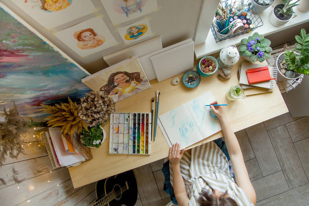 The girl artist sits at her workplace and paints portraits in watercolors. Finished works are hanging on the wall. She has a gray cat in her arms. On the table are brushes, paints, paper, tea and flowers. - Photo, Image
