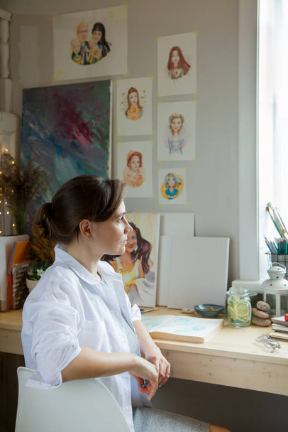 The girl artist sits at her workplace and paints portraits in watercolors. Finished works are hanging on the wall. She has a gray cat in her arms. On the table are brushes, paints, paper, tea and flowers. - Foto, afbeelding