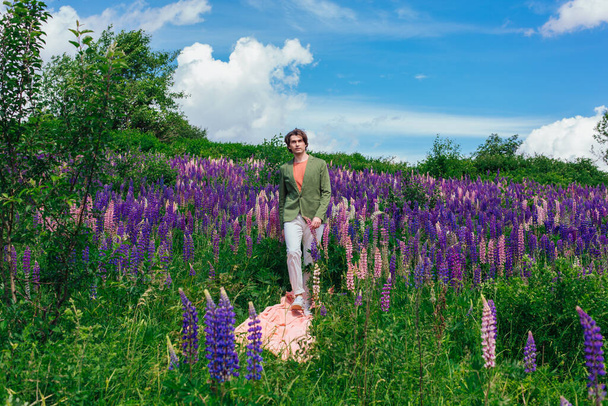 Tall handsome man in a green jacket walking on a pink cloth in lupine flowers field and enjoing the beauty of nature. Man surrounded by purple and pink lupines. - Foto, Bild