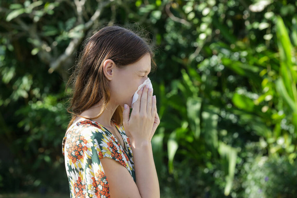 Сезонная аллергия. Молодая девушка, сморкающаяся и чихающая в тканях в парке.  - Фото, изображение