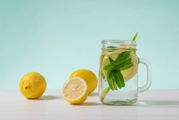 Большая кружка холодной воды с лимоном и мятой на белом столе на голубом фоне. - Фото, изображение