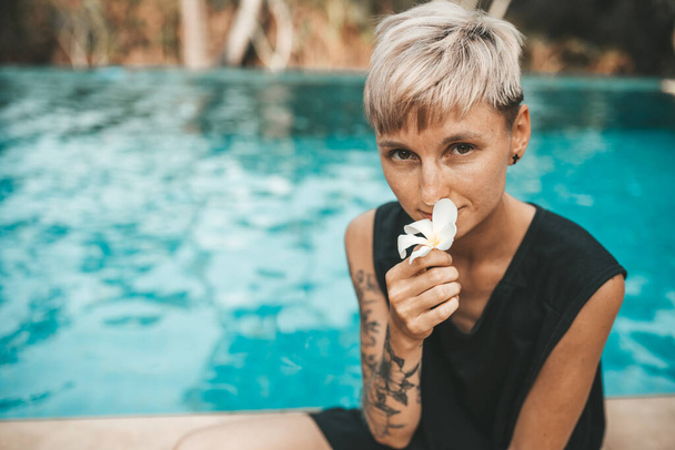 Woman blonde sitting at poolside and sniffing a flower frangipani - Fotoğraf, Görsel