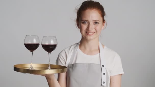 Pretty smiling waitress in apron holding tray with red wine over white background - Footage, Video