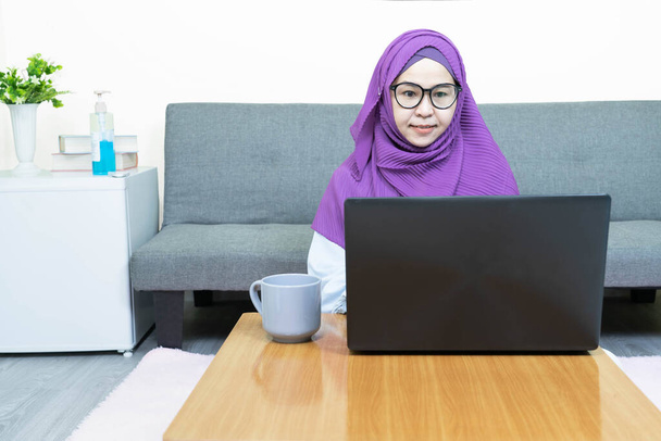 Young beautiful asian muslim woman in traditional dress wearing glasses  using laptop in living room at home,female chatting in social network, shopping or working online, looking at computer screen - Φωτογραφία, εικόνα