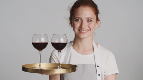 Όμορφη χαμογελαστή σερβιτόρα σε ποδιά κρατώντας δίσκο με κόκκινο κρασί κοιτάζοντας χαρούμενα στην κάμερα πάνω από γκρι φόντο - Πλάνα, βίντεο