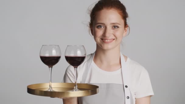 Attraktive lächelnde Kellnerin in Schürze mit Tablett und Wein, die vor weißem Hintergrund selbstbewusst in die Kamera blickt - Filmmaterial, Video