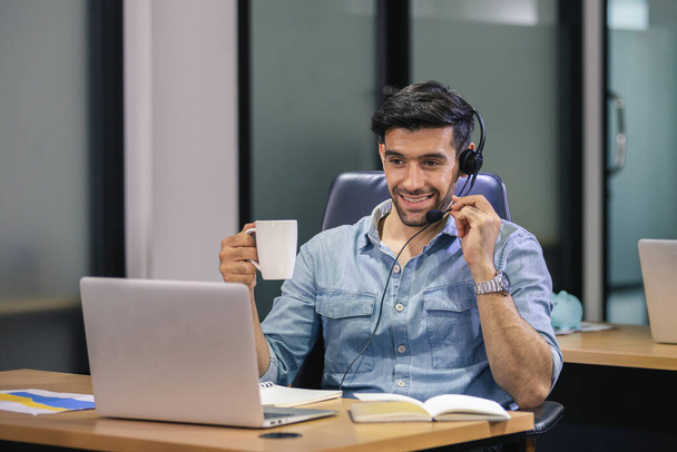 pohledný muž mluvící mikrofon se sluchátky vypadající notebook drží šálek kávy u stolu práce v kanceláři, on-line konference, práce nové normální. - Fotografie, Obrázek