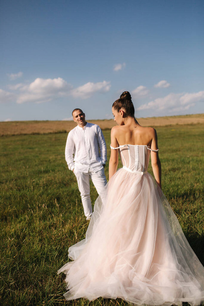 Gorgeous bride with handsome groom walkin in the field after wedding ceremony. Newlyweds posing to photographer - Photo, Image