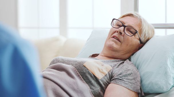 Κοντινό πλάνο μιας γριάς άρρωστης γυναίκας ξαπλωμένης στο κρεβάτι να μιλάει με έναν άντρα βοηθό - Φωτογραφία, εικόνα