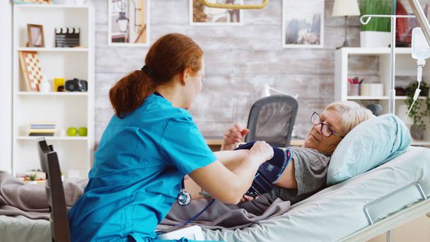 Enfermera midiendo la presión arterial de anciana enferma acostada en cama de hospital
 - Foto, Imagen