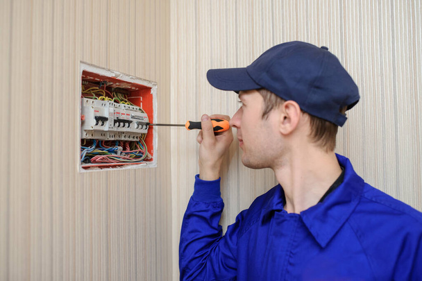 boční pohled na mladého elektrotechnika v modrém celkovém rozebrání elektrického panelu s pojistkami v pouzdře - Fotografie, Obrázek