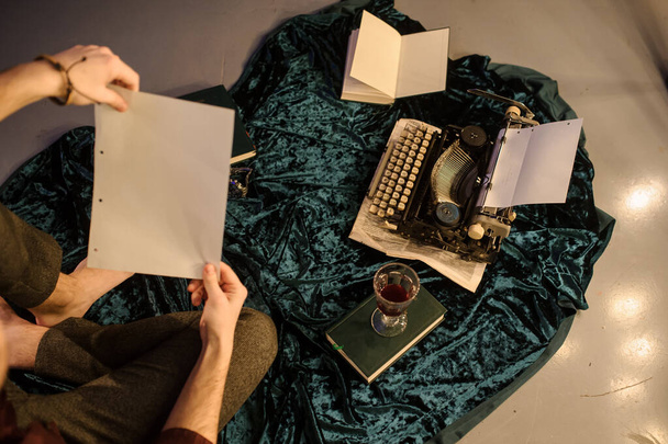 vista dall'alto dell'uomo con un pezzo di carta tra le mani seduto vicino a una macchina da scrivere vintage su un tessuto di velluto blu scuro sul pavimento con fogli di carta e libri arround - Foto, immagini