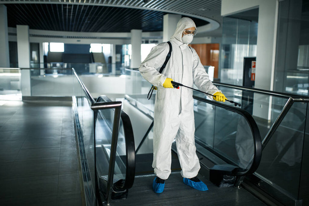Trabalhador de desinfecção vestindo um terno de proteção limpa profissionalmente uma escada rolante em um centro de negócios vazio. Um homem equipado com roupas antibacterianas esteriliza shopping center. Conceito de Covid
 - Foto, Imagem