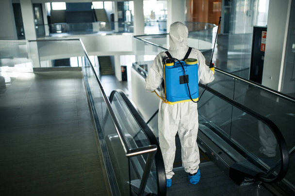 Ένας υπάλληλος απολύμανσης που φοράει προστατευτική στολή καθαρίζει επαγγελματικά μια κυλιόμενη σκάλα σε ένα άδειο επιχειρηματικό κέντρο. Ένας άντρας εξοπλισμένος με αντιβακτηριακά ρούχα αποστειρώνει το εμπορικό κέντρο. Έννοια Covid - Φωτογραφία, εικόνα