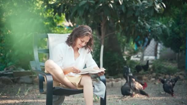 A young cheerful woman sits in a chair in the garden and reads a book - Video