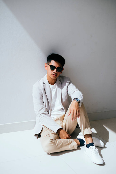 Un hombre de traje blanco con gafas de sol actúa como un modelo en la pared blanca con luz y sombra
. - Foto, Imagen