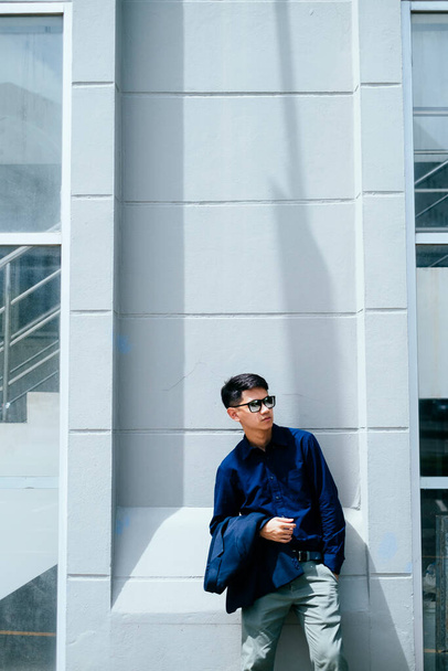 Ein Mann in dunkelblauem Hemd mit Sonnenbrille steht im Sommer im Sonnenlicht vor dem Gebäude. - Foto, Bild
