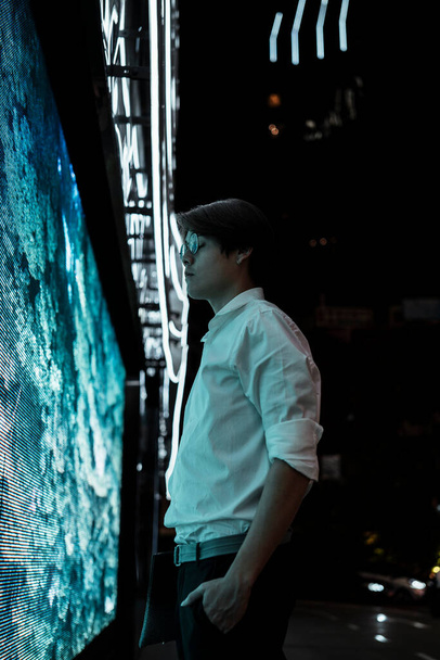 Homem de cabelo preto em camisa branca e óculos de pé na tela LED em frente ao shopping na cidade à noite
. - Foto, Imagem