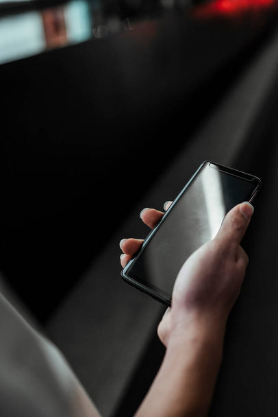 Zugeschnittenes Bild des Smartphones in der Hand des Mannes, während er mit dunklem Boden als Hintergrund geht. - Foto, Bild