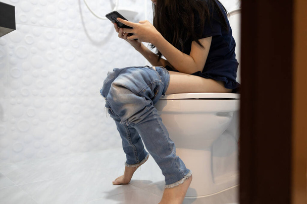 Азиатская девочка с запором, женщина, играющая по телефону в онлайн-игру в ванной комнате или зависимость от социальных сетей, люди, сидящие на унитазе в течение длительного времени может вызвать геморрой, образ жизни - Фото, изображение