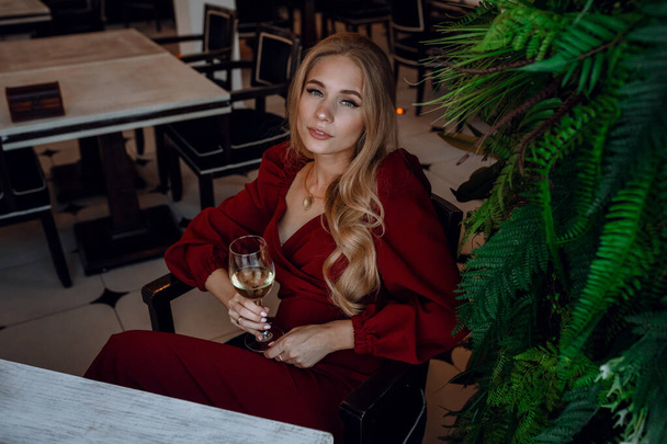 カフェやレストランで白ワインを飲む女性です。セクシーな若いブロンドの女の子。お祝いとかお酒とかスタイルとかファッショナブルなライフスタイルとか。肖像画の女の子で赤ドレスでワイングラス - 写真・画像
