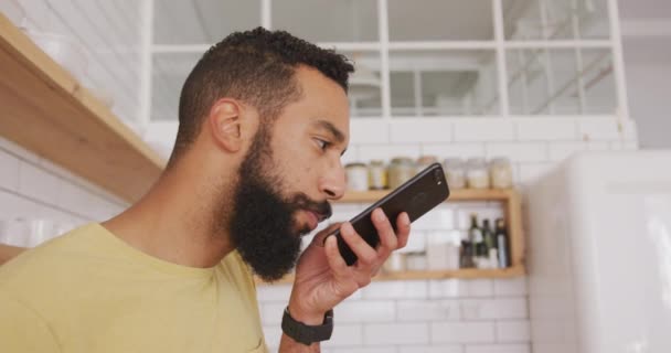 Mixed-race man relaxing at home at the weekend, standing in a kitchen, by a table, talking on a  smartphone. Social distancing in quarantine lock down - Imágenes, Vídeo