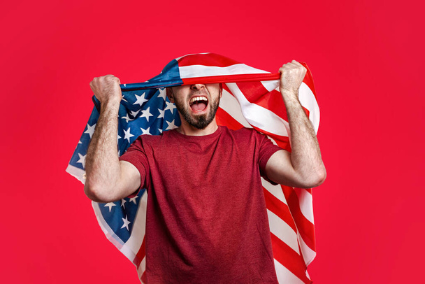Белый мужчина с американским флагом на голове поднимает руки и кричит. Красный фон. Концепция патриотизма, власти, свободы и спорта болельщиков. - Фото, изображение