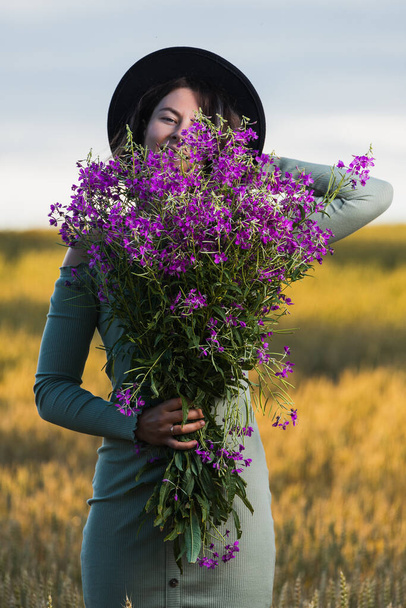 Hermosa mujer en un lindo vestido azul, sombrero negro y un enorme ramo de flores silvestres moradas se ríe al aire libre en un prado durante la puesta del sol. Concepto de caminar estilo de vida
 - Foto, imagen