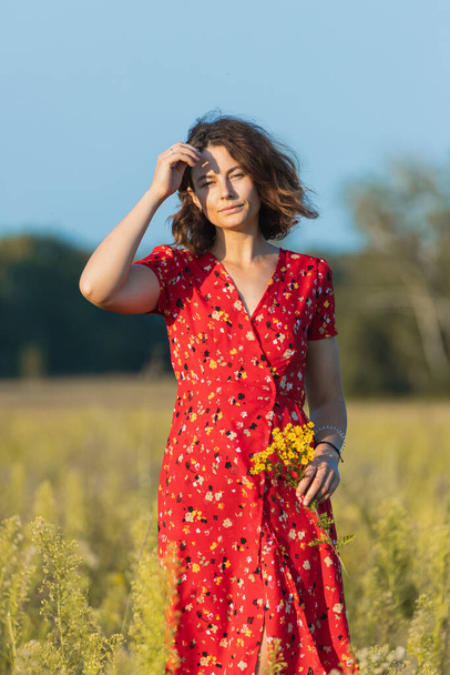 Portrait of a beautiful young woman   in red romantic dress with yellow flowers walking on the field , enjoying the day, on a sunny summer day. The concept of the unity of the child and nature, peaceful mood, environmental life - Photo, Image
