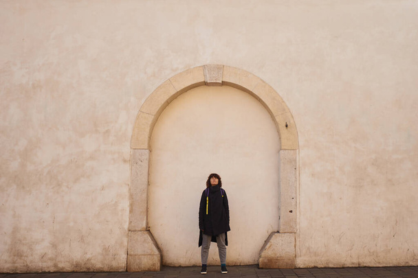 Nuori kaunis brunette turisti nainen kihara tukka ja musta takki seisoo iso seinä tausta ja etsii suoraan kameraan. Elämäntapa ja matkustaminen käsite. - Valokuva, kuva