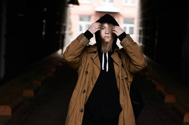 City portrait of attractive hipster girl in trendy black hoodie and brown trench. Beautiful carefree woman in posing near dark wall with city background. Lifestyle concept. - Foto, immagini