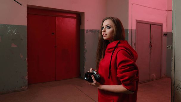 A beautiful girl of 20-25 years old in a red sweatshirt holds a mirrorless camera in an underground room.  - Foto, Imagen