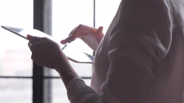Zijaanzicht close-up van de handen van een vrouw drukt op een touch screen werken op een tablet staat in de buurt van grote ramen in een helder kantoor  - Video