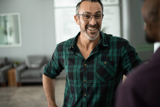 Зрелый бизнесмен разговаривает с коллегой и смеется, стоя вместе в современном офисе - Фото, изображение