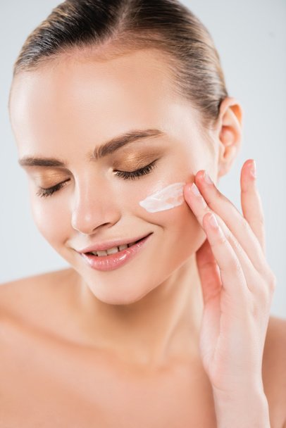 cheerful woman with closed eyes applying face cream isolated on grey  - Foto, imagen