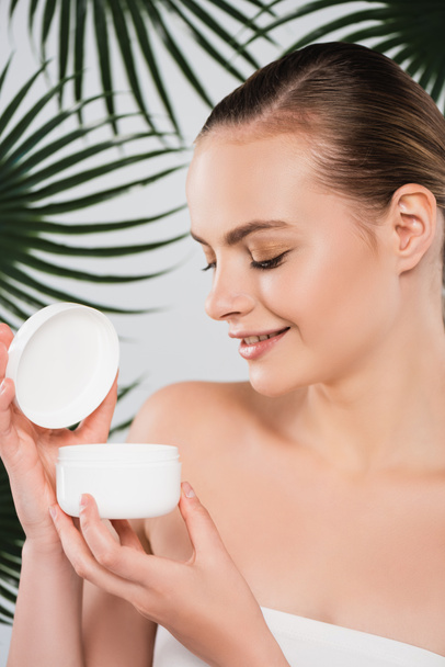 happy woman looking at container with face cream near palm leaves on white - Photo, Image