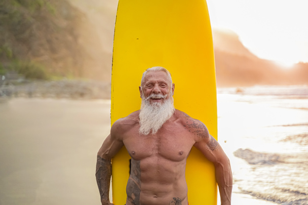 Idősebb, aki napnyugtakor szörfözik. Sportos szakállas férfi edzés szörfdeszkával a parton. Idősek egészséges életmód és extrém sport koncepció. Kép - Fotó, kép
