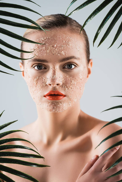 naked woman with makeup and scrub on face looking at camera near palm leaves on white - Fotografie, Obrázek