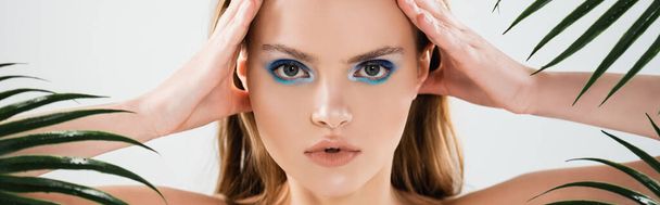 horizontal crop of beautiful woman with blue eye shadow looking at camera near palm leaves on white - Foto, imagen