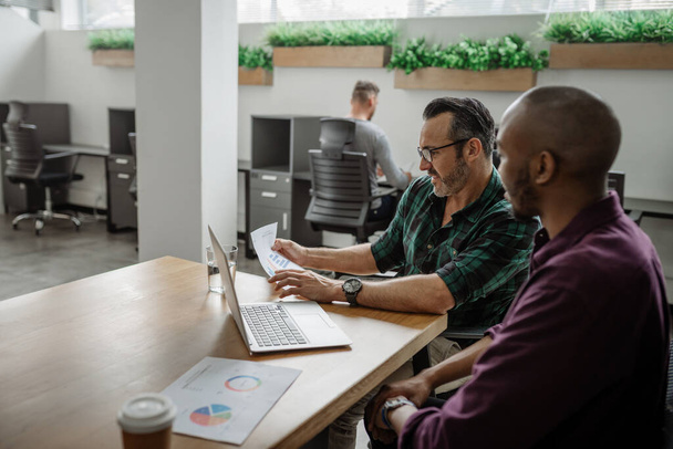 Dois empresários diversos que trabalham em um laptop e revisam documentos enquanto se sentam em uma mesa em um escritório - Foto, Imagem